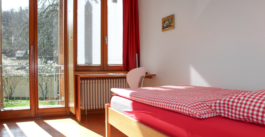 Foto: Schlicht eingerichtetes Hotelzimmer mit Einzelbett und Blick durch die gläserne Terrassentür ins Grüne.