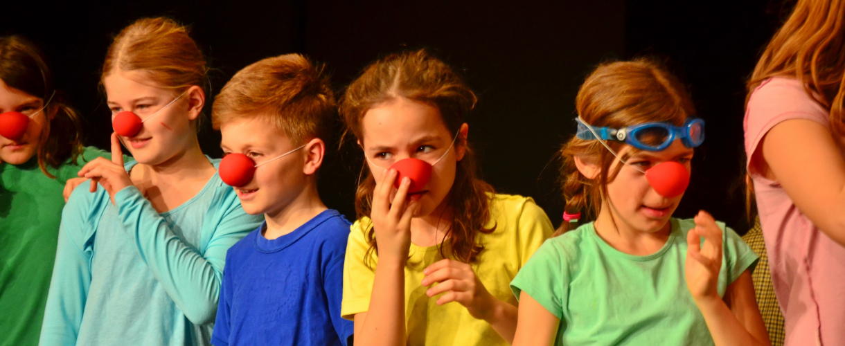 Foto: Kinder stehen nebeneinander auf einer Bühne und tragen rote Clownsnasen; sie wirken konzentriert und bereiten sich gemeinsam auf einen Auftritt vor.