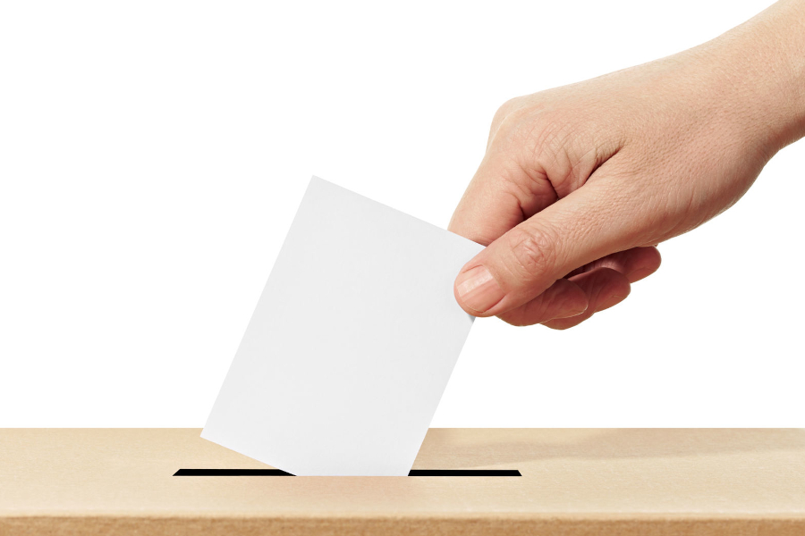 Link zur Seite Demokratiebildung (Foto: Eine Hand hält einen weißen Zettel, um ihn in den Schlitz einer Wahlurne zu werfen.)