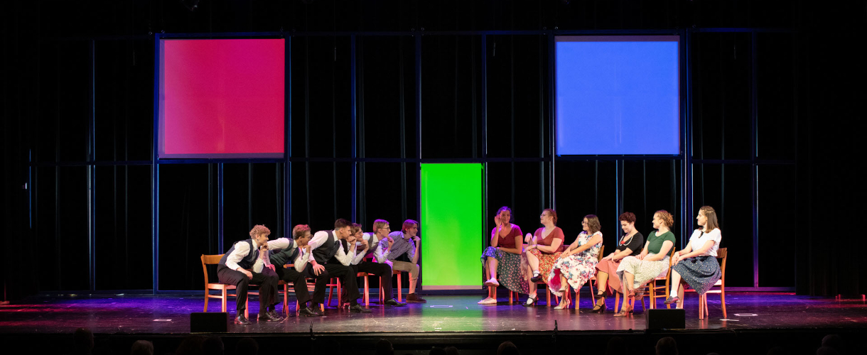Foto einer Szene: Auf einer Bühne stehen zwei Stuhlreihen in V-Form, auf denen rechts Mädchen in Kleidern aus den dreißiger Jahren und links Jungen mit Anzügen aus dreißiger Jahren sitzen und sich anschauen.