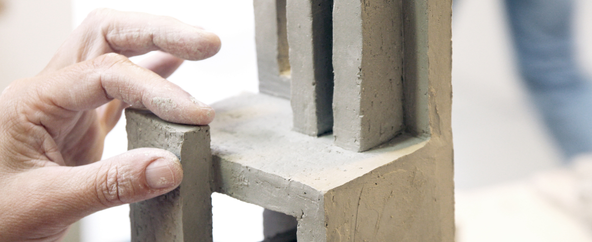 Foto: Eine Hand formt im Kunstunterricht eine architektonische Skulptur aus Ton mit klaren geometrischen Formen.