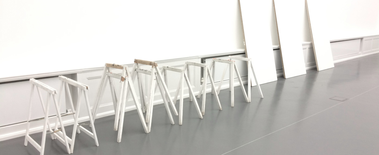 Foto: Weiße Holzgestelle und Tischplatten lehnen an einer Wand in einem hellen, leeren Ausstellungsraum – ein Bild für kreative Freiräume und bevorstehende künstlerische Prozesse.