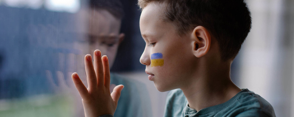 Symbolbild für: Das Thema Krieg im Unterricht (Foto: Junge, der eine ukrainische Flagge auf die Wange gemalt hat, blickt traurig aus dem Fenster.)