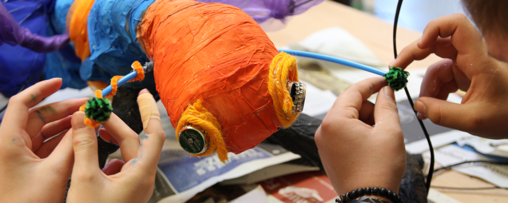 Foto: Eine bunt bemalte Pappmachèfigur wird von zwei Paar Kinderhänden gemeinsam bearbeitet.