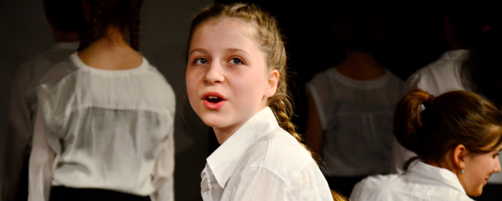 Foto von einer Theaterszene mit drei sitzenden Personen. Ein Mädchen mit Zopf und weißer Bluse dreht sich zum Publikum um und spricht mit ausdrucksvollem Gesicht.