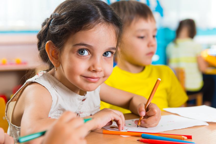 Symbolbild für Grundschulen (Foto: eine Grundschülerin sitzt malend an einem Tisch im Klassenzimmer und schaut aufgeweckt in die Kamera; neben ihr sitzt ein Grund-schüler, der vor sich hinblickt; im Hintergrund sitzt eine weitere Grundschülerin mit dem Rücken zum Betrachter)