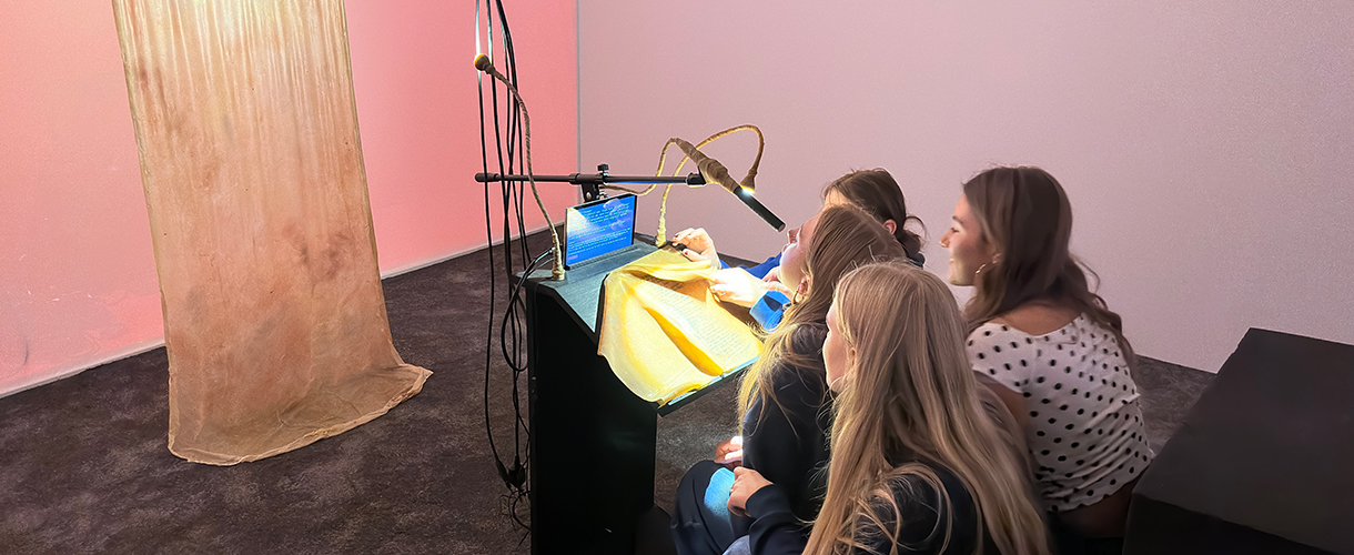 Foto: Vier Schülerinnen betrachten eine Kunstinstallation aus Textil in einem Raum mit rosa Hintergrundbeleuchtung und interagieren mit dem Kunstwerk durch ein Mikrophon und einen Monitor.