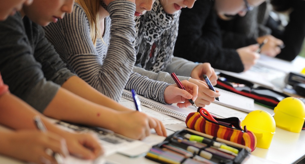Link; Symbolbild Besondere Förderbedarfe (Foto: Schüler schreiben konzentriert in Hefte; Federmäppchen und Stifte liegen auf den Tischen im Klassenraum.)