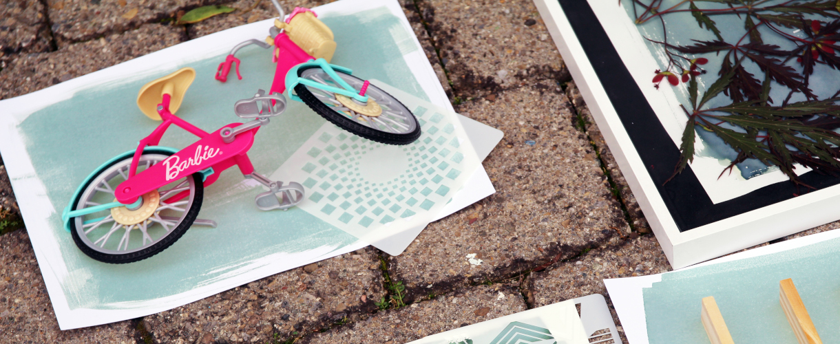 Foto: Auf einem Pflasterboden liegen künstlerische Drucke, darunter ein Bild mit einem pinken Barbie-Fahrrad sowie eine gerahmte Cyanotypie mit Blättern.