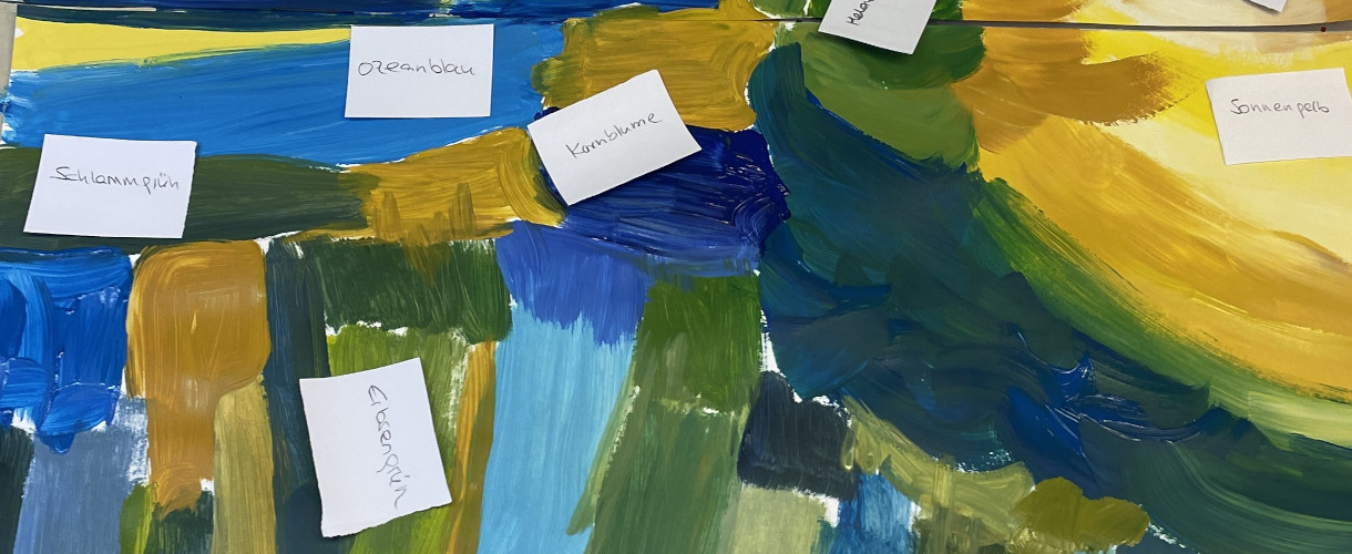 Foto: Ein abstrakt gemaltes Bild in Blau-, Grün- und Gelbtönen mit handbeschrifteten Papierzetteln, wie „Ozeanblau“, „Kornblume“, „Schlammgrün“ und „Sonnengelb“, die einzelne Farbflächen benennen.