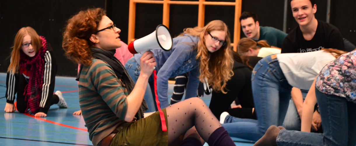Foto aus einem Theaterworkshop: Mittig sitzt eine Person auf dem Fußboden und spricht in eine Flüstertüte, während um sie herum viele Personen auf dem Boden knien und zu ihr schauen.