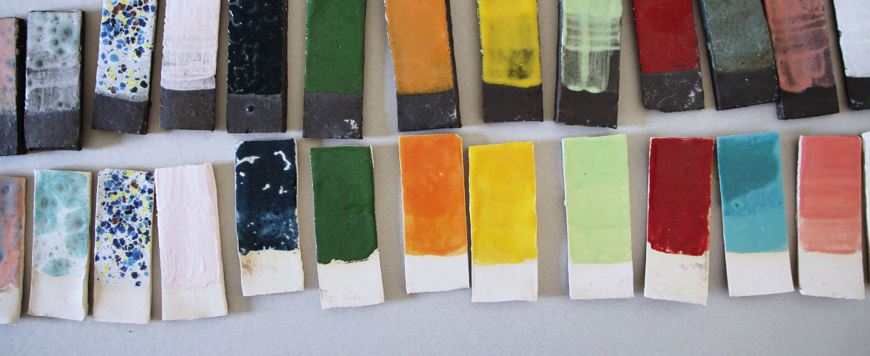 Foto: Das Bild zeigt zwei Reihen rechteckiger Keramik-Testplättchen mit unterschiedlich farbigen Glasurproben, von Grau, Weiß und Grün über Orange und Geld bi zu Rot, Türkis und Rosa. 