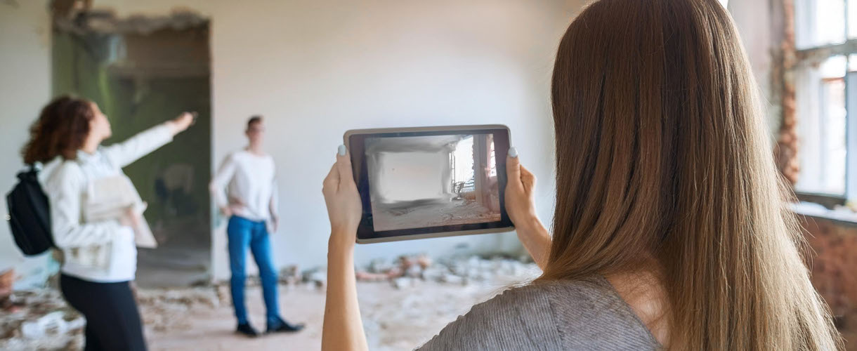 Foto: Eine Frau filmt mit einem Tablet einen teils renovierten Raum, während zwei Personen im Hintergrund Details besprechen.
