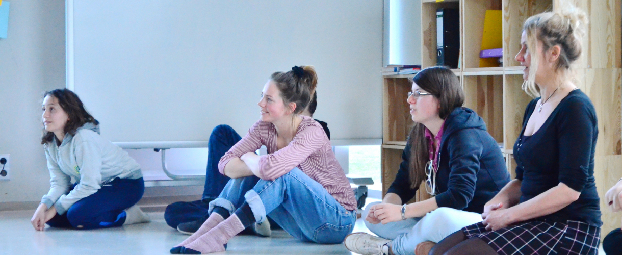 Foto aus einem Theaterworkshop: Teilnehmerinnen eines Workshops sitzen auf dem Fußboden eines Klassenzimmers und schauen zur linken Seite.