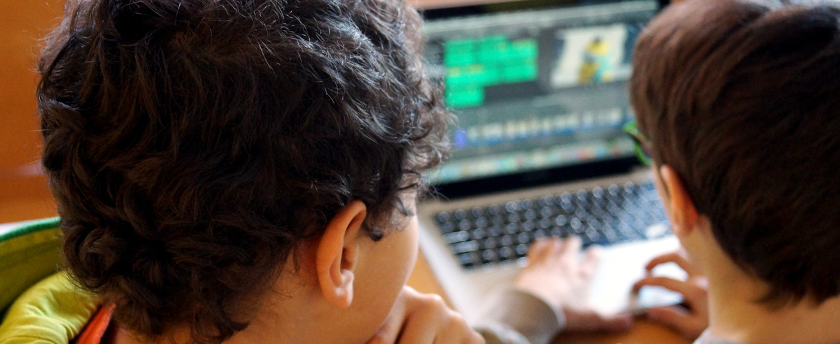 Foto: Zwei Kinder sitzen von hinten gesehen dicht nebeneinander vor einem Laptop und arbeiten gemeinsam an einer Videobearbeitungssoftware. 