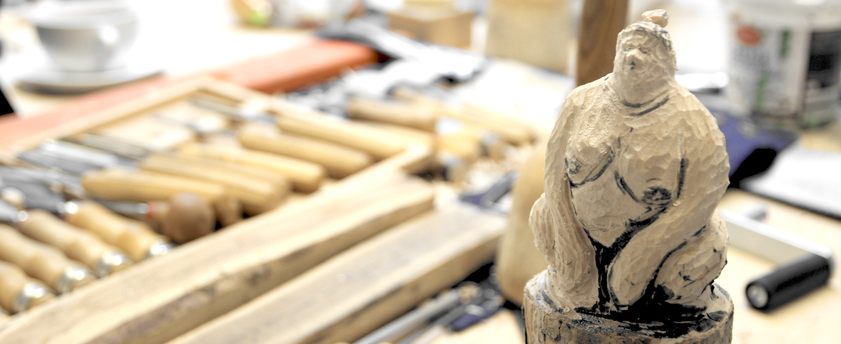 Foto von voluminöser hockender Figur aus grob geschnitztem Holz, eventuell Sumo-Ringer. Im Hintergrund sind unscharf einige Schnitzwerkzeuge mit Holzgriffen zu erkennen.