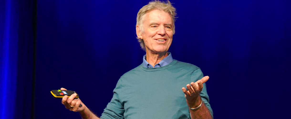 Prof. John Hattie hält einen Vortrag bei der Fachtagung: „Lernen sichtbar machen – Chancen pädagogischen Handelns in Kita und Grundschule“ am 23.09.2025 in Heilbronn (Foto: Zu sehen ist der Oberkörper John Hatties, der einen grünen Pullover und darunter ein blaues Hemd trägt. Er lächelt und hat beide Arme angehoben, mit den Handflächen nach oben. In der rechten Hand hält er eine Fernbedienung.)