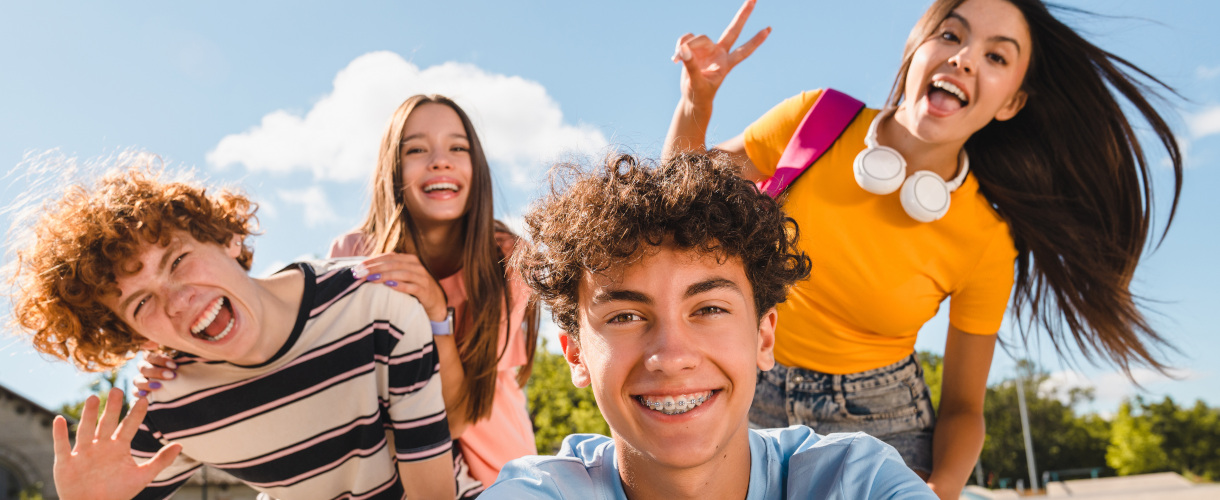 Link zur Aktuelles-Meldung: Kick-off Dialekt und Du – das Webportal für sprachliche Vielfalt (25. September 2026, 11 bis 12 Uhr, Anmeldeschluss: 20. September 2026) (Foto: Vier Jugendliche vor blauem Himmel, zwei Jungen und zwei Mädchen, lächeln und winken begeistert in die Kamera.)