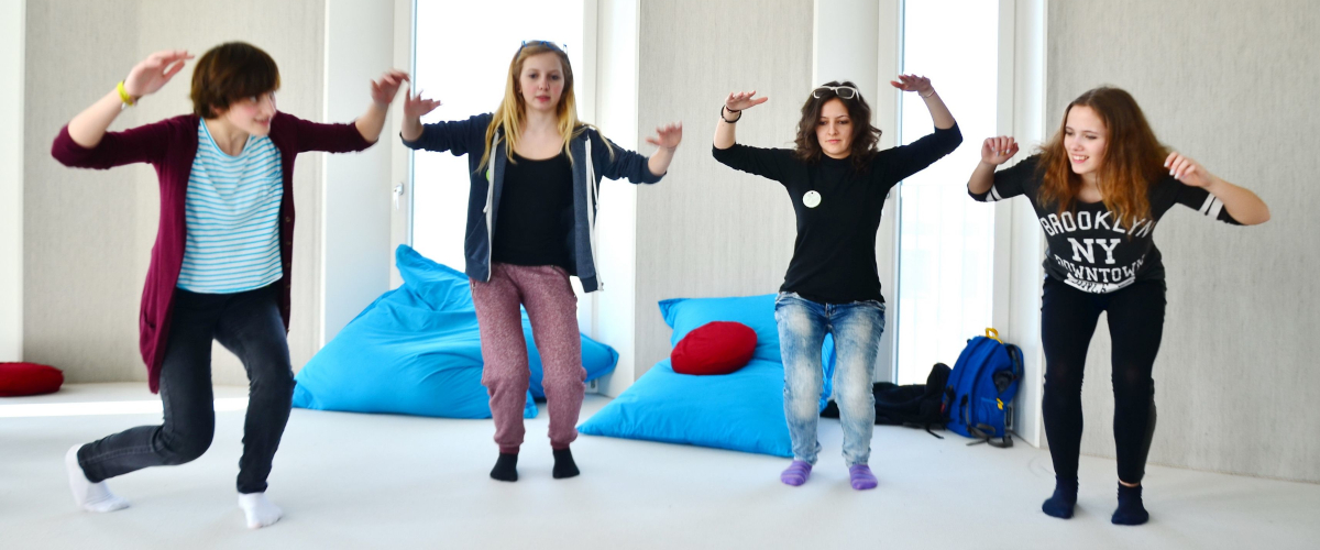 Foto aus einem Theaterworkshop: Vier Personen stehen vor einem weißen Hintergrund mit großen türkisen Sitzsäcken und gehen langsam in die Knie, während sie ihre Arme auf Schulterhöhe halten.