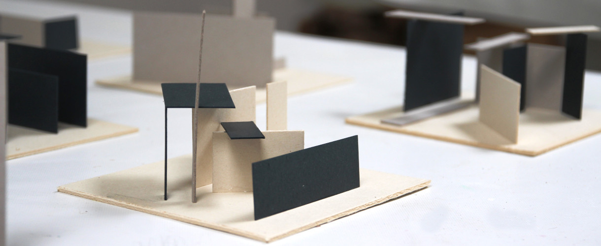 Foto: Mehrere kleine Architekturmodelle aus weißem Karton und schwarzen Papierflächen stehen auf einem Tisch. 
