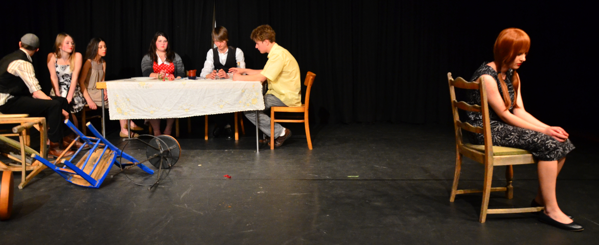 Foto einer Szene: Vor einem schwarzen Hintergrund befindet sich auf der linken Seite ein Tisch, an dem viele Menschen sitzen, sowie umgeworfene Stühle am Boden; auf der rechten Seite steht ein einzelner Stuhl, auf dem eine Person sitzt, die dem Tisch den Rücken zukehrt und die Hände in den Schoss gelegt hat.