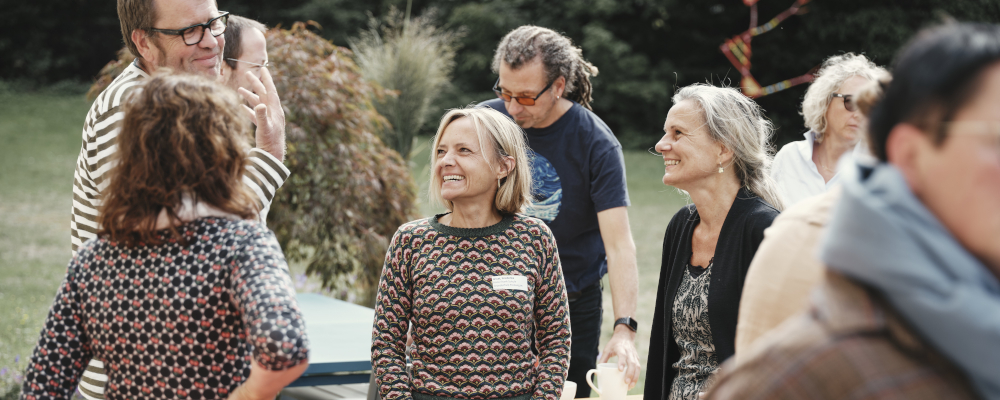 Foto: Stehende Menschengruppe bei einer Kaffeepause auf einer Terrasse. Vier Personen unterhalten sich lächelnd miteinander.