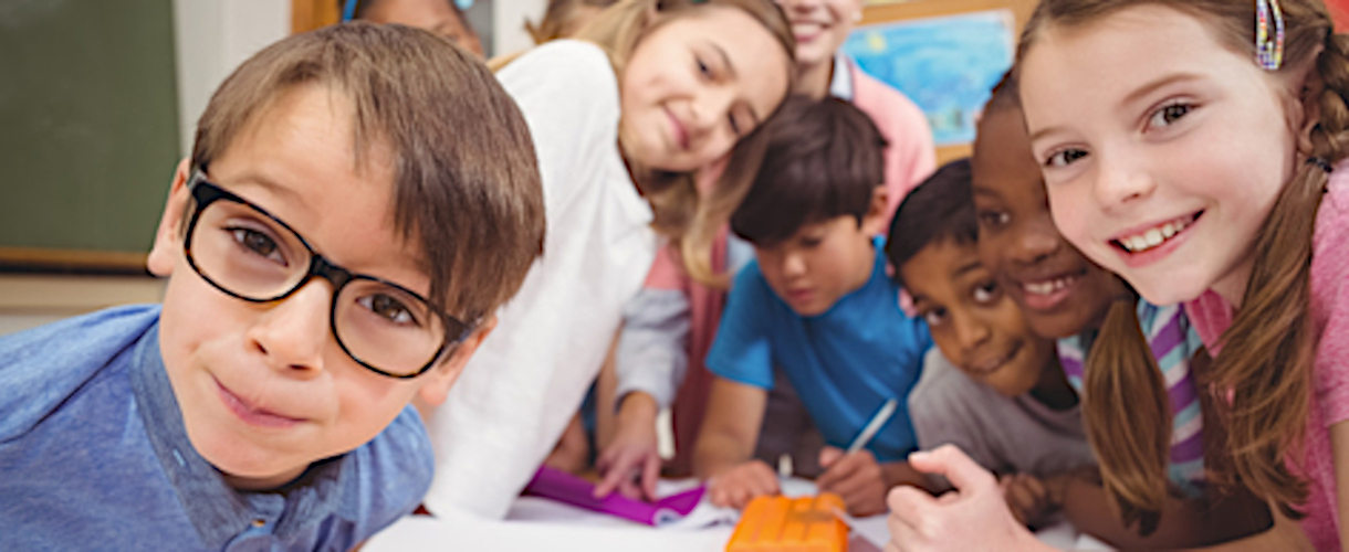 Link zur ZSL-Aktuelles-Meldung: Zertifikatskurs „Starke BASIS! Mathematik Grundschule“ (Bad Wildbad, 14. bis 15. Dezember 2026) (Foto: Mehrere Kinder, Jungen und Mädchen, sitzen oder lehnen im Klassenzimmer an einem Gruppenarbeitstisch. Sie schauen lächelnd in die Kamera. Im Hintergrund ist eine Lehrerin zu erahnen.)