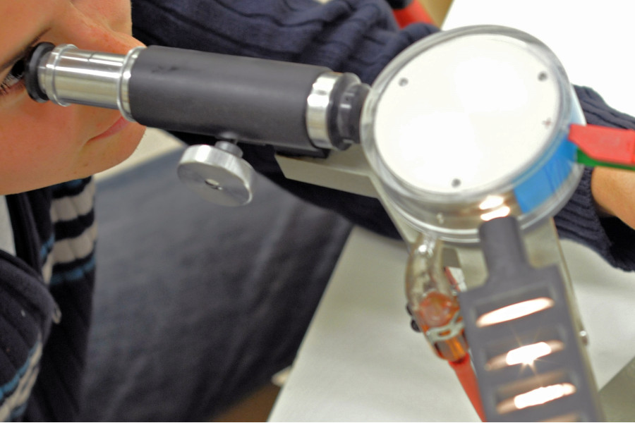 Link zur ZSL-Seite „Physik“; Foto: Ein Schüler sitzt vor einem physikalischen Experiment und schaut durch ein Okular.