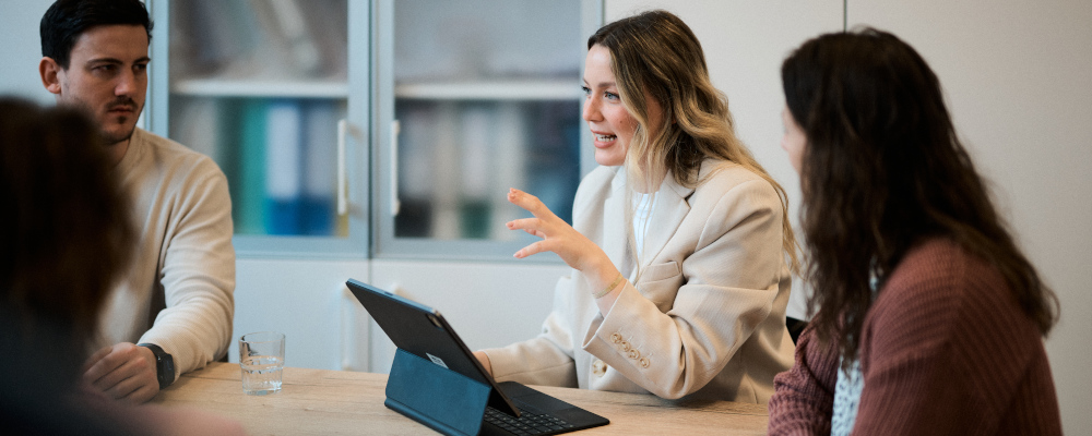 Symbolbild für Schulaufsicht (Foto: Vier Personen sitzen an einem Tisch. Im Zentrum ist eine Frau im Kostüm, die vor einem Laptop sitzt und den anderen mit der linken Hand gestikulierend etwas erläutert. Links von ihr sitzen ein Mann und rechts von ihr, weiter im Vordergrund, eine Frau, die ihr zuhören. Die vierte Person links vorne ist nur als Schatten erahnbar.)