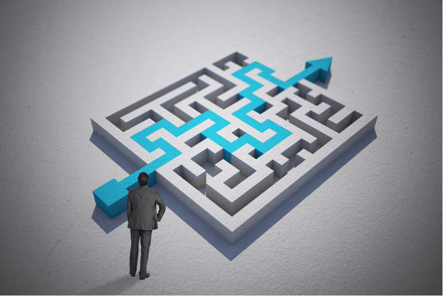 Link zur ZSL-Seite Förderkonzepte; Symbolbild für Förderkonzepte (Grafik: Das Bild zeigt eine stilisierte 3D-Labyrinth-Grafik auf hellem Hintergrund. Am unteren Rand steht eine Person in einem dunklen Anzug mit dem Rücken zur Betrachtung und blickt auf das Labyrinth. Ein hervorgehobener Weg in türkisblauer Farbe führt vom Eingang unten links durch das Labyrinth und endet oben rechts mit einem Pfeil, der aus dem Labyrinth herauszeigt.)