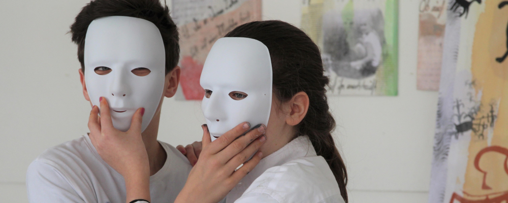 Foto: Zwei in Malhemden gekleidete Jugendliche befinden sich in einem Atelier und halten sich gegenseitig eine weiße Theatermaske vors Gesicht.