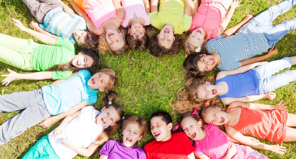 Link; Symbolbild Gesundheitsförderung (Foto: 14 Kinder in bunter Kleidung liegen mit den Köpfen zugewandt rücklings auf einem grünen Rasen)