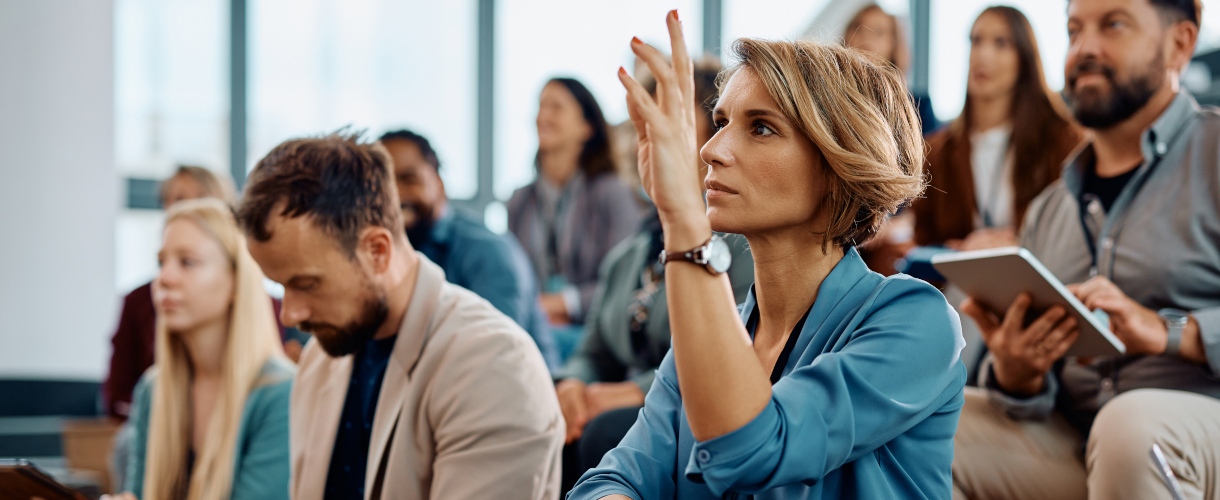 Link zur Aktuelles-Meldung: Fortbildungsreihe für neu bestellte Schulleitungen: Der Prozess der Ziel- und Leistungsvereinbarungen (Foto: Mehrere Personen mittleren Alters sitzen in einem Hörsaal. Manche machen sich Notizen. Im Fokus des Bildes ist eine Frau zu sehen, die die Hand hebt, um eine Frage zu stellen oder einen Kommentar abzugeben.)