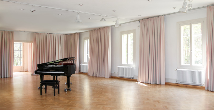 Foto von einem lichtdurchfluteten großen Raum mit Parkettboden und hellen Vorhängen. In der Mitte steht ein schwarzer Konzertflügel. 