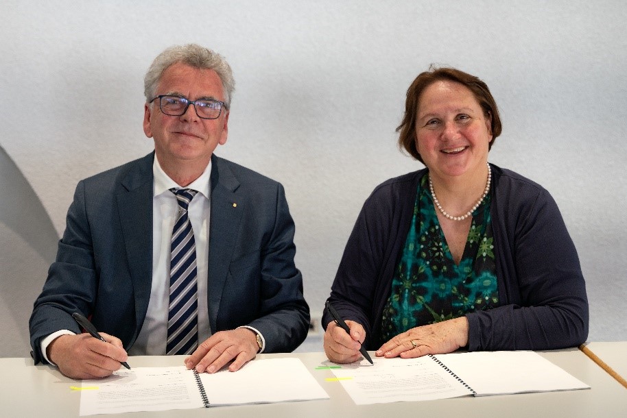 Foto: Uwe Bleich, Vorstand der Hector Stiftungen, in Anzug mit gestreifter Krawatte, sitzt (aus Betrachterperspektive) links an einem Tisch. Theresa Schopper, baden-württembergische Kultusministerin, mit grünschwarzgemustertem Oberteil und schwarzer Jacke sitzt rechts von ihm. Beide lächeln in die Kamera. Sie halten beide einen Stift in der Hand und haben jeweils zwei Blätter vor sich liegen. Sie sind dabei, die Vereinbarung zur Verlängerung ihrer Zusammenarbeit mit den Hector Kinderakademien zu unterzeichnen.