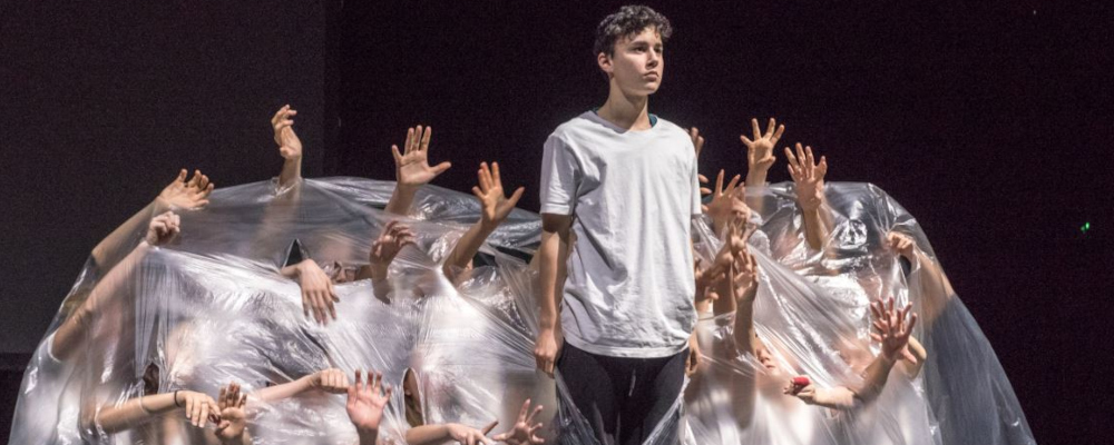 Theaterszene (Foto: Vor schwarzem Hintergrund ist eine große durchsichtige Folie zu sehen, unter der Menschen knien oder hocken. Man kann ihre Gesichter nicht erkennen. Sie strecken ihre Arme und Hände durch Risse in der Folie nach außen. Zentral sichtbar steht eine junge Person, die sich durch einen Schlitz in der Folie ganz aufgerichtet hat.)