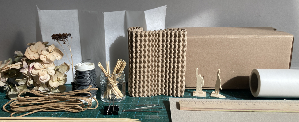 Foto: Materialsammlung für Architektur-Workshops: getrocknete Blüten, Papierbahnen, Drahtrollen, Kordel, Holzstäbchen im Glas, Wellpappe, Kartonbox, kleine Modell-figuren, Schneidernadeln, Lineal und Schneidematte auf einem Arbeitstisch.