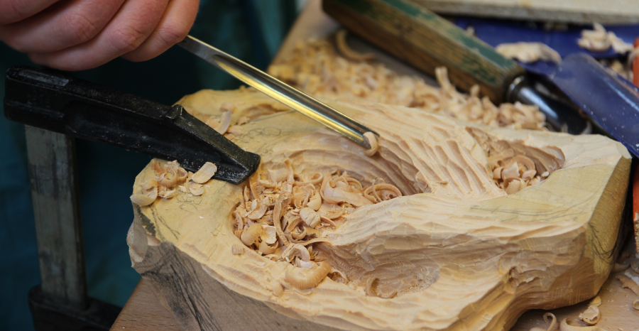 Link zur ZSL-Seite „Lehrkräftefortbildung Bildende Kunst“; Fotografie von einem Werkstück aus Holz, das gerade von einer Hand mit einem Werkzeug bearbeitet wird.