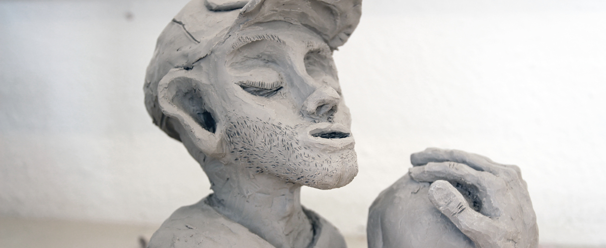 Foto: im Kunstunterricht modellierte Tonfigur eines jungen Mannes mit geschlossenen Augen, Bartstoppeln und einer Mütze, der eine große Kugel in den Händen hält