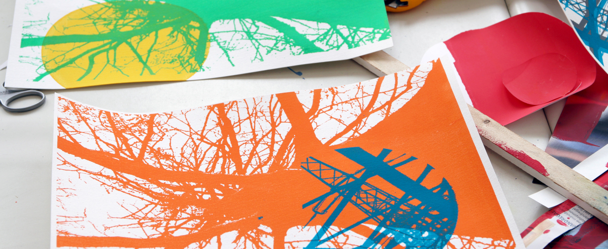 Foto: zwei farbige Siebdrucke mit stilisierten Baum- und Architekturmotiven in Orange-Blau und Grün-Gelb liegen auf einem Tisch, umgeben von Schere, Papier und weiteren Gestaltungsmaterialien