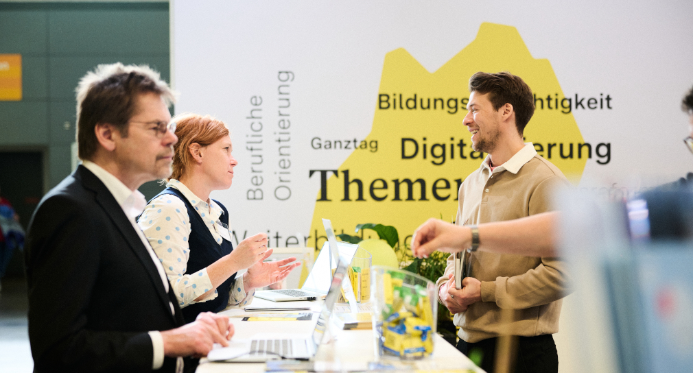 Fotografie: Austausch am ZSL-Stand, Blick auf den Tresen des Messestandes. Drei Personen im Austausch