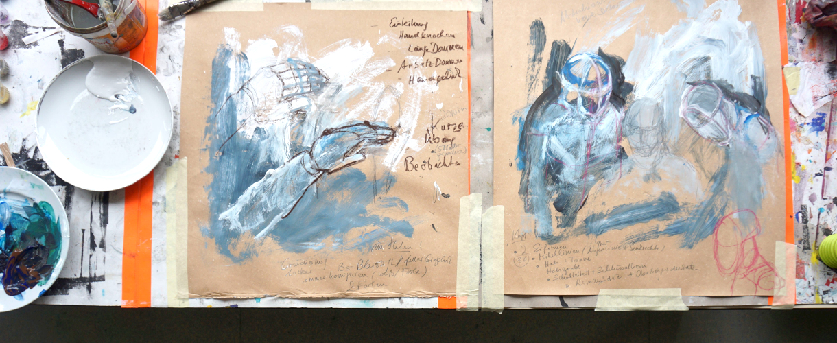 Foto: Zwei expressive Figuren- und Handstudien in Acryl und Buntstift auf braunem Papier, umgeben von Malutensilien auf einem Arbeitstisch.