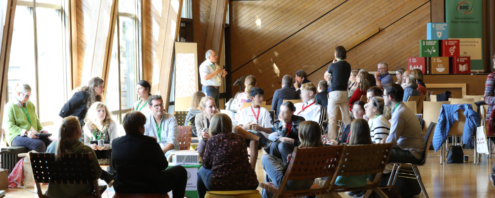 BNE-Netzwerktreffen (Foto: Um die dreißig Personen sitzen in mehreren Stuhlkreisen beisammen. Eine große Gruppe im Vordergrund tauscht sich im Gespräch aus. Eine große Gruppe im Hintergrund hört dem Vortrag eines Mannes zu.)