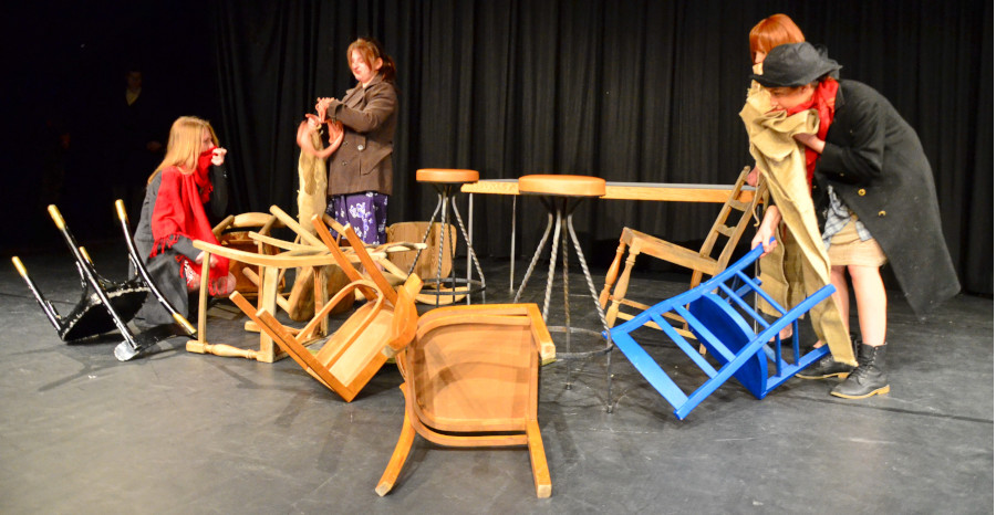 Link zur ZSL-Seite „Lehrkräftefortbildung Theater“; Foto einer Schultheaterszene: Auf einer Theaterbühne mit einem Tisch und mehreren umgefallenen Stühlen agieren vier Personen in Kostümen.