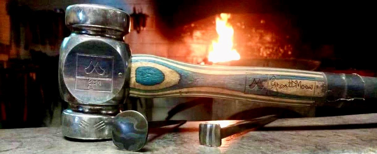 Foto: Ein Schmiedehammer mit Holzgriff liegt auf einem Amboss. Daneben steht ein kleines Metallstück, im Hintergrund brennt das Feuer einer Schmiede.