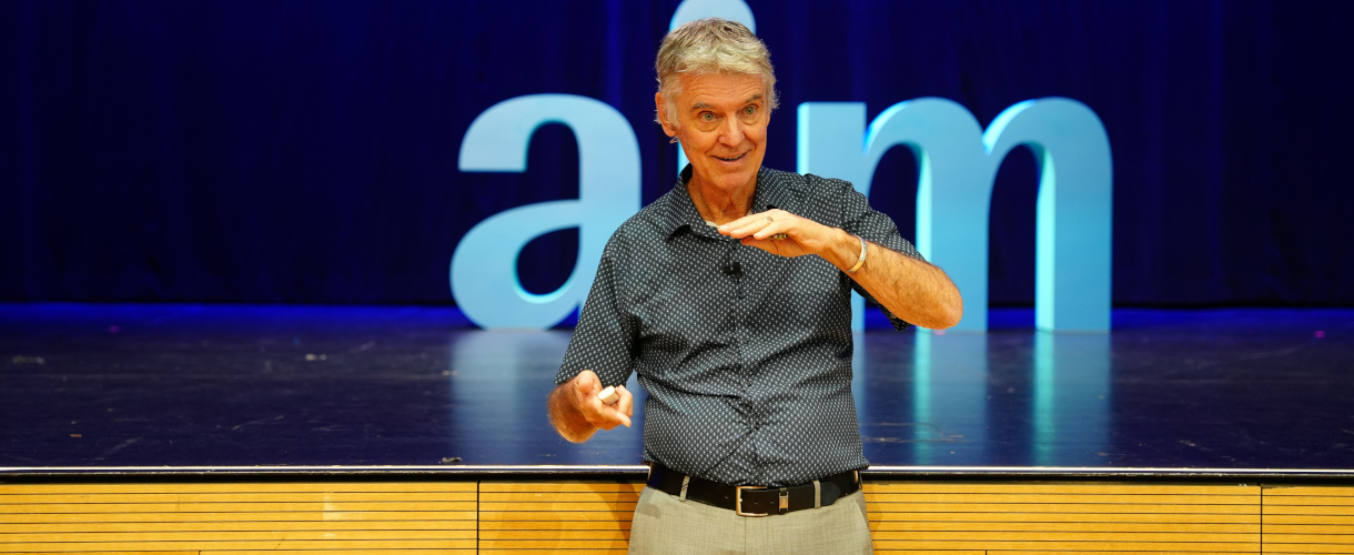 Prof. John Hattie hält Keynote bei der Masterclass Mindframes of Leaders (Heilbronn, 22.09.2025) (Foto: John Hattie, der ein dunkles Hemd trägt, steht vor der Bühne, auf der das aim-Symbol zu sehen ist, und unterstreicht seinen Vortrag mit ausladenden Armbewegungen.)