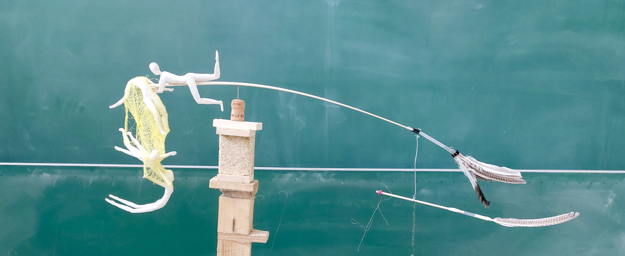 Foto: Mobile aus Holz, Draht, Federn und Figuren vor grüner Schultafel, das Bewegung und Balance thematisiert.