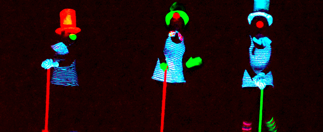 Foto: drei clownhafte Personen in Neonfarbe beleuchtet von Schwarzlicht