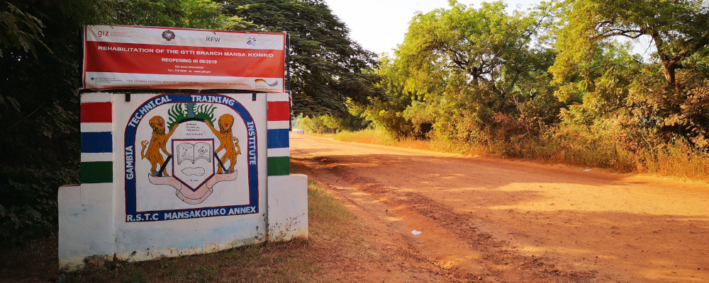 GTTI-Ausbildungsbehörde in Mansa Kongo - Projektpartner des ZSL (Foto: trockene, von Bäumen und Büschen umrandete Straße in Afrika; links neben der Straße steht ein großes Schild mit dem Titel „Rehabilitation of the GTTI Branch Mansa Konko Reopening in 08/2019. Darunter ist ein Wappen mit zwei Löwen, die links und rechts auf ihren Hinterläufen neben einem aufgeklappten Buch und einer Erdnuss stehen. Um das Wappen herum steht: Gambia Technical Training Institute R. S. T. C Mansakonko Annex)